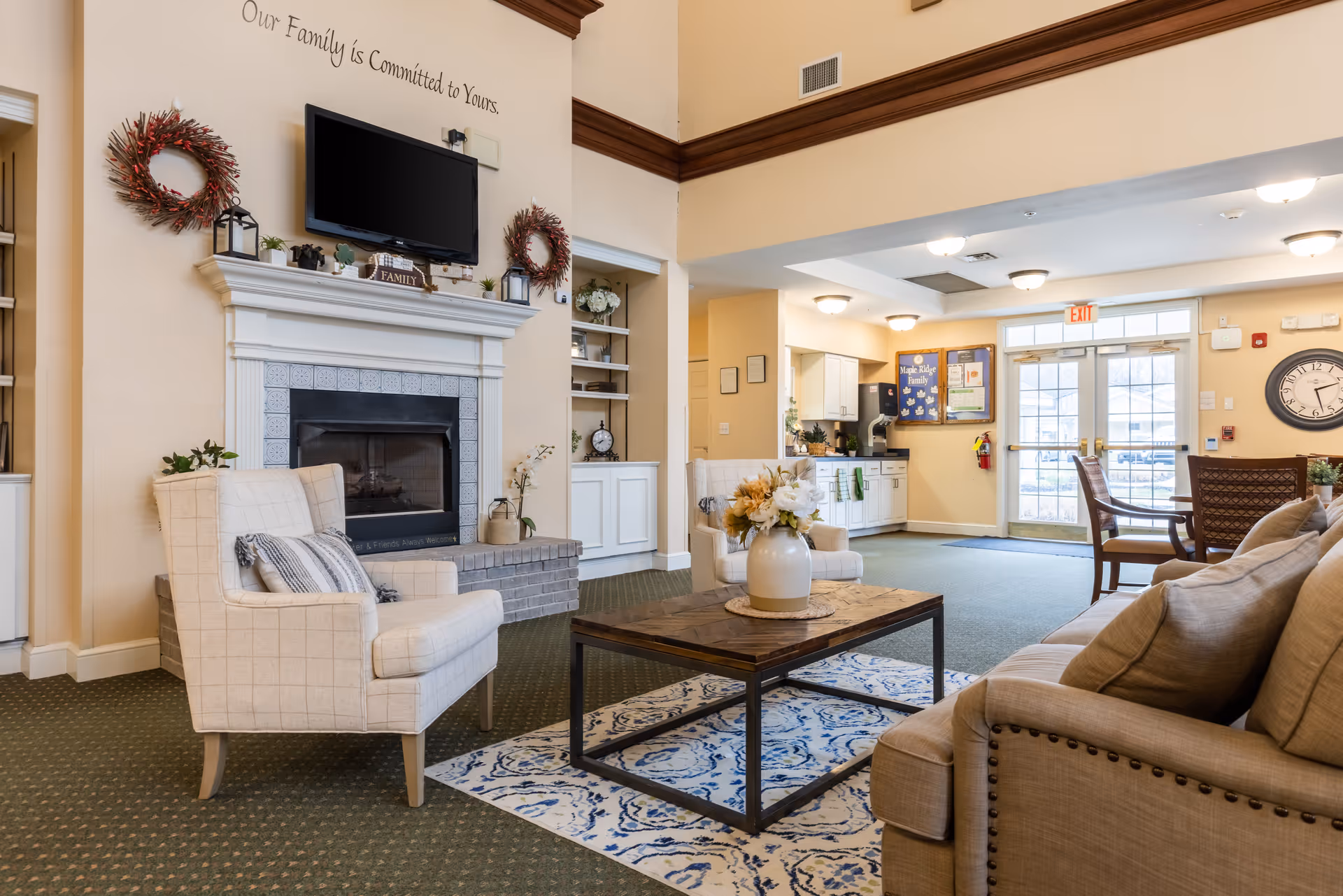A cozy senior living common area with beige walls and green carpet. The room features a white fireplace with a mounted flat-screen TV above it, flanked by two decorative wreaths. There are two beige armchairs and a beige sofa arranged around a wooden coffee table with a floral centerpiece on a patterned rug. In the background, there is a kitchenette area and glass double doors leading outside. A large clock is mounted on the wall near the doors.