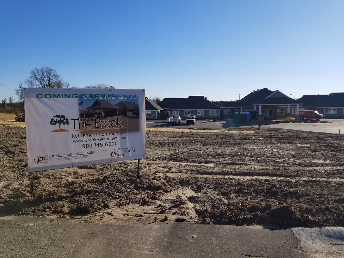 A construction site with a large banner for The Brook retirement community in front of a low one-story building under a clear blue sky.