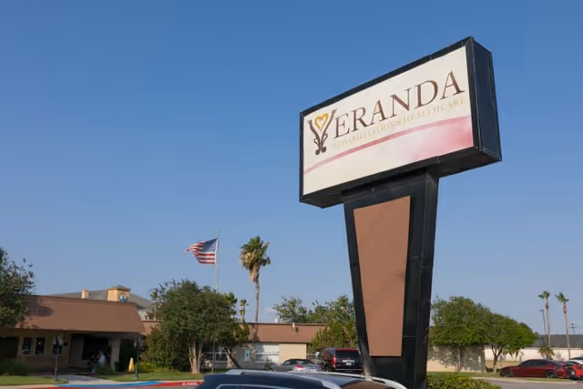Outdoor view of Veranda Rehabilitation And Healthcare facility sign with the building, trees, parked cars, and a clear blue sky in the background.