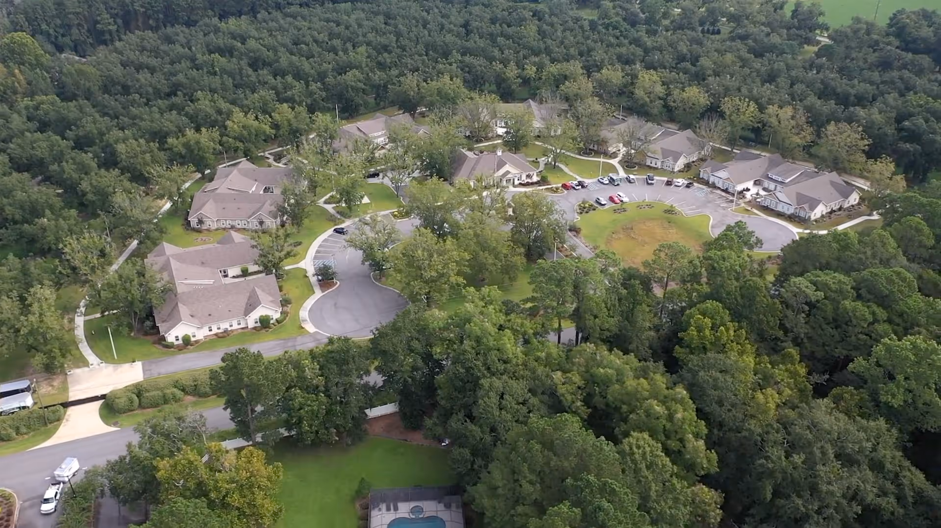 Aerial view of The Orchard At Stone Creek senior living facility surrounded by dense trees and greenery, showing multiple single-story buildings arranged in a circular layout with parking areas and pathways connecting them.