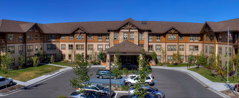 Front exterior view of Luther Park at Sandpoint, a large multi-story senior living facility with a covered entrance, multiple windows, and a parking lot with several cars. The building features a combination of stone and wood siding with a brown roof, surrounded by landscaped greenery and trees under a clear blue sky.