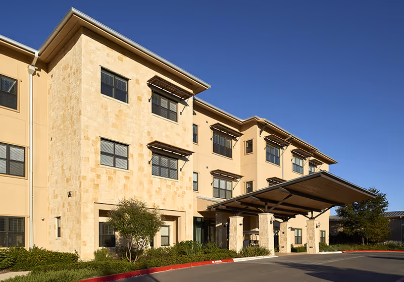 Exterior view of a three-story assisted living facility building with beige stone and stucco walls, multiple windows with awnings, a covered entrance supported by stone pillars, and landscaping with bushes and trees under a clear blue sky.