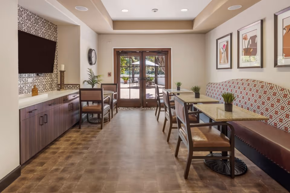 A bright dining area with multiple tables and chairs arranged along a patterned bench seat on the right side. The left side features a long wooden cabinet with a decorative tiled backsplash and a mounted flat-screen TV. A clock and potted plants are also visible. Double glass doors at the end of the room lead to an outdoor patio with umbrellas and greenery.