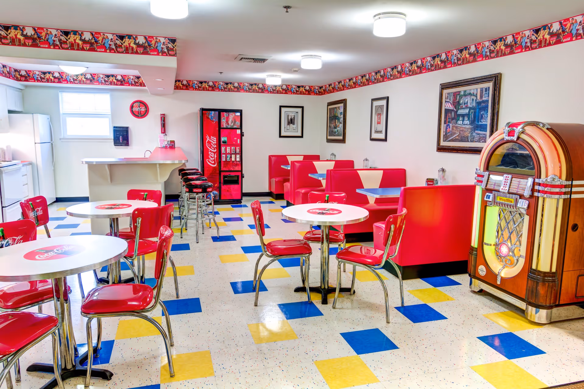 A retro-themed dining area with red and white Coca-Cola branded tables and chairs, red booth seating along the wall, a vintage jukebox, a Coca-Cola vending machine, and a small kitchen area with a refrigerator and stove. The floor has a colorful pattern with blue, yellow, and white tiles, and the walls are decorated with framed pictures and a Coca-Cola border near the ceiling.