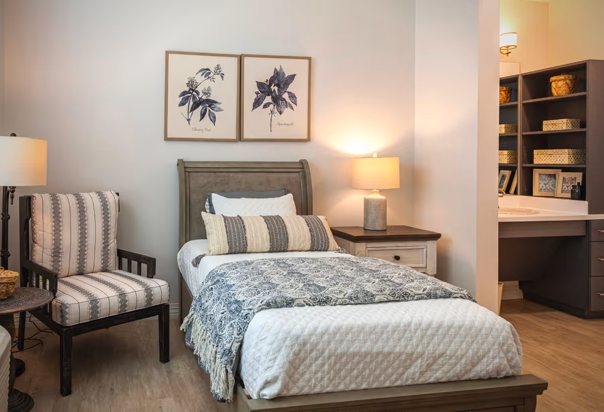 A cozy bedroom featuring a single bed with white and patterned bedding, a striped armchair, two framed botanical prints on the wall, a wooden nightstand with a lamp, and a vanity area with shelves holding baskets and framed photos.