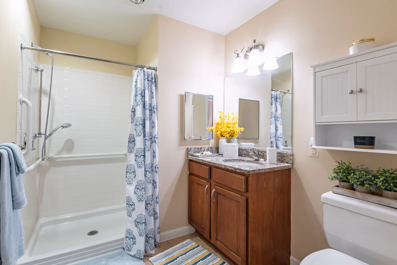 A clean and well-lit bathroom featuring a walk-in shower with a white and blue patterned curtain, a wooden vanity with a granite countertop, a large mirror with three light fixtures above, and a white cabinet mounted on the wall above the toilet. The bathroom is decorated with yellow flowers on the vanity and green plants on the toilet tank.