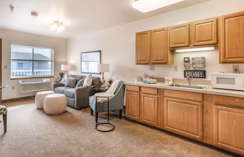 A cozy living area in a senior living facility with a gray sofa, two white ottomans, a blue armchair with a side table, and a kitchen area with wooden cabinets, a sink, and a microwave. A window with blinds and an air conditioning unit is visible in the background.