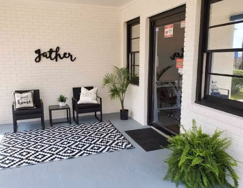 A covered patio area with two black chairs, each with decorative pillows, a small black side table with a plant, a black and white geometric patterned rug, potted plants, and a white brick wall with the word 'gather' mounted on it. There is a glass door and windows with black frames on the right side.