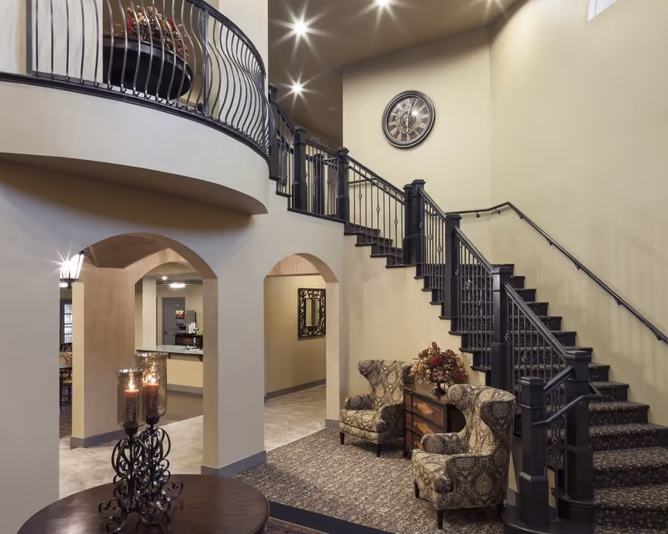Interior view of a senior living facility lobby area featuring a staircase with black railings, two patterned armchairs, a wooden side table with a floral arrangement, a round table with decorative candle holders, and a large wall clock. The space has beige walls and arched doorways leading to other rooms.