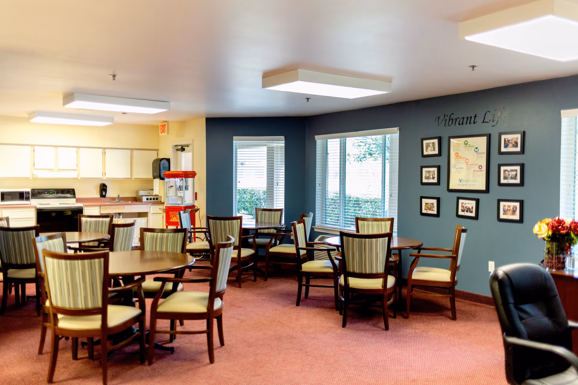 A bright dining area in a senior living facility with several round wooden tables surrounded by cushioned chairs. The room has large windows with blinds, a blue accent wall with framed pictures and the words 'Vibrant Life' displayed. In the background, there is a kitchenette with white cabinets, a stove, microwave, and a red popcorn machine. A black office chair and a vase with flowers are visible on the right side.