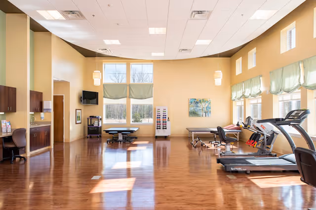 A spacious rehabilitation center room with wooden floors and large windows letting in natural light. The room contains exercise equipment including treadmills and therapy tables. There are chairs, cabinets, and a wall-mounted TV, with light yellow walls and green window valances.