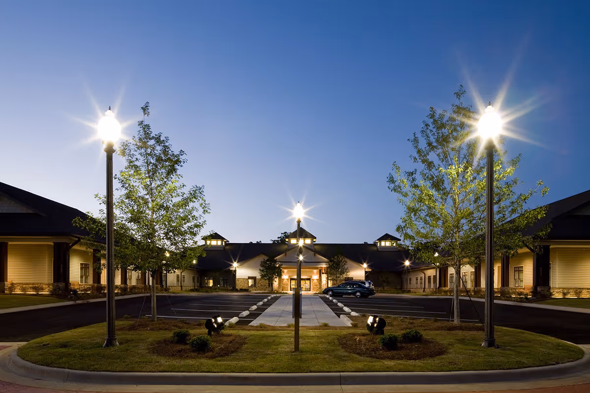 Exterior view of Oaks on Parkwood senior living facility at dusk, showing a well-lit entrance with street lamps, trees, and a parking lot with a few cars.