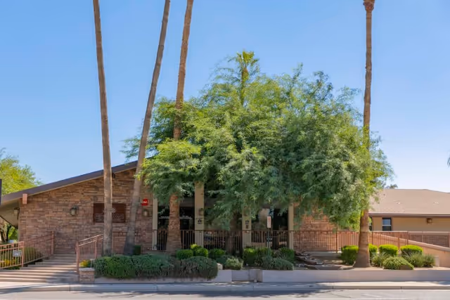 Exterior view of Heritage Court Post Acute Of Scottsdale building with stone facade, surrounded by tall palm trees and lush green bushes under a clear blue sky.