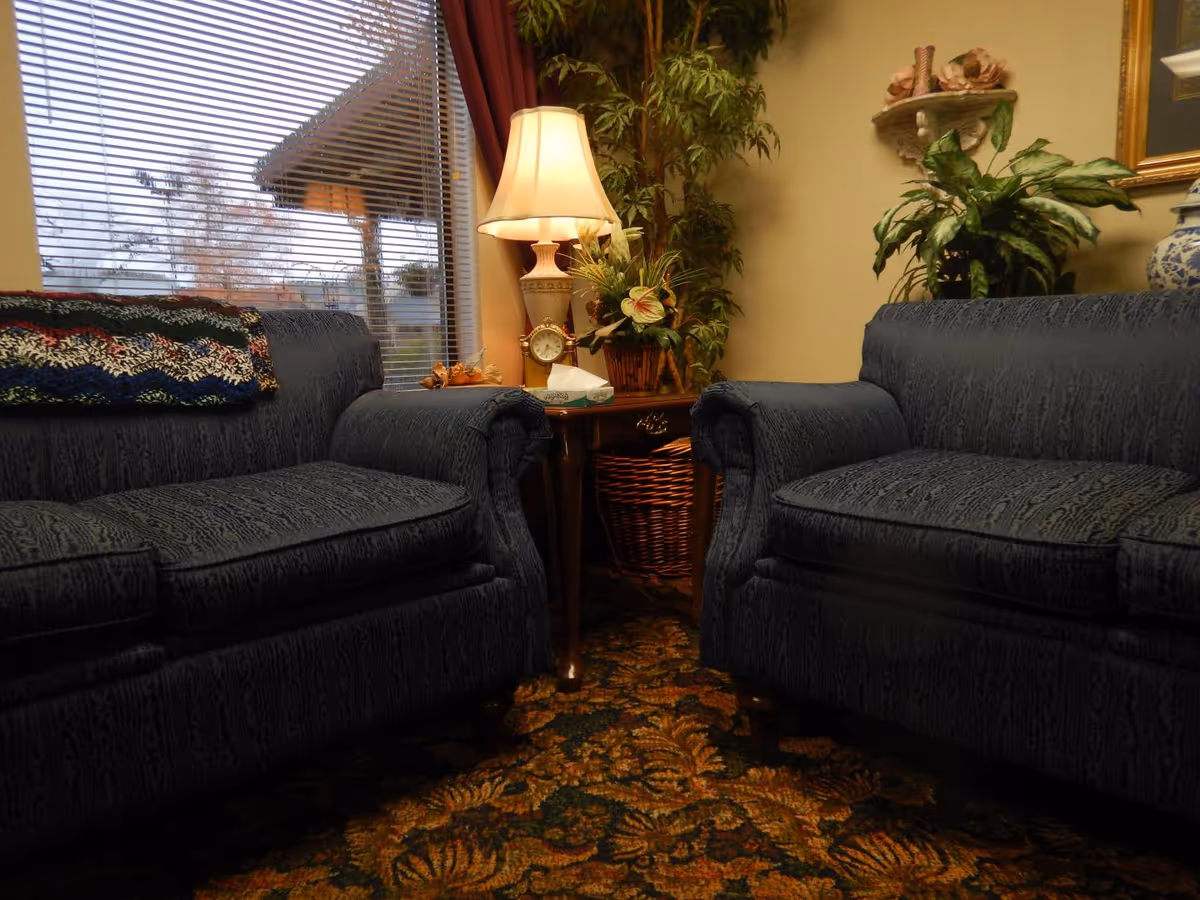 A cozy living room area with two dark blue upholstered armchairs facing each other. Between them is a wooden side table with a decorative lamp, a clock, a tissue box, and a flower arrangement. Behind the table is a tall green plant, and a window with blinds and red curtains is visible on the left side. The floor is covered with a patterned carpet.