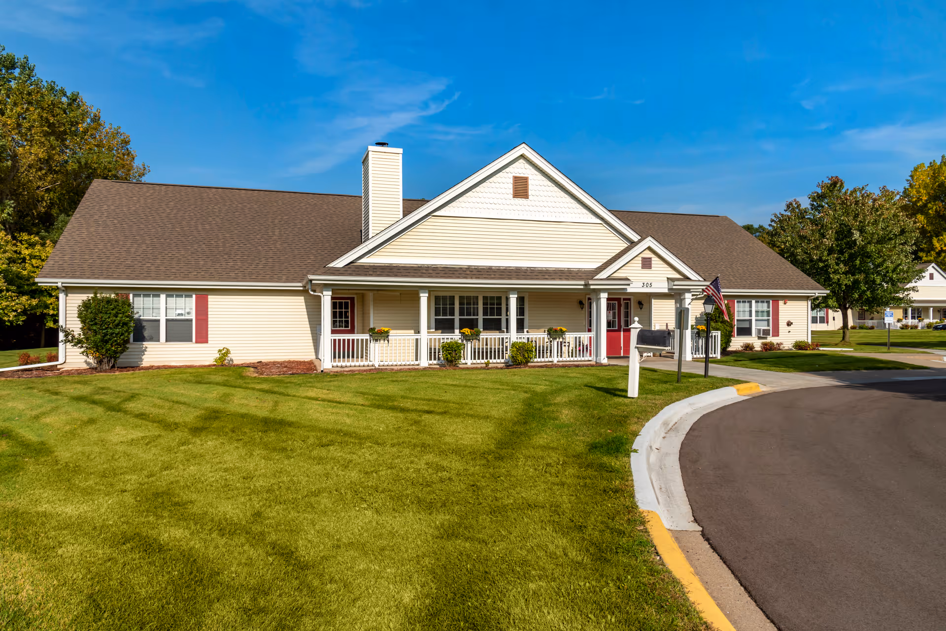Exterior view of a single-story residential building with beige siding, a brown roof, and red doors and window shutters. The building has a covered porch with white railings and flower pots. There is a well-maintained green lawn in front and a curved paved driveway to the right. Trees and another similar building are visible in the background under a clear blue sky.