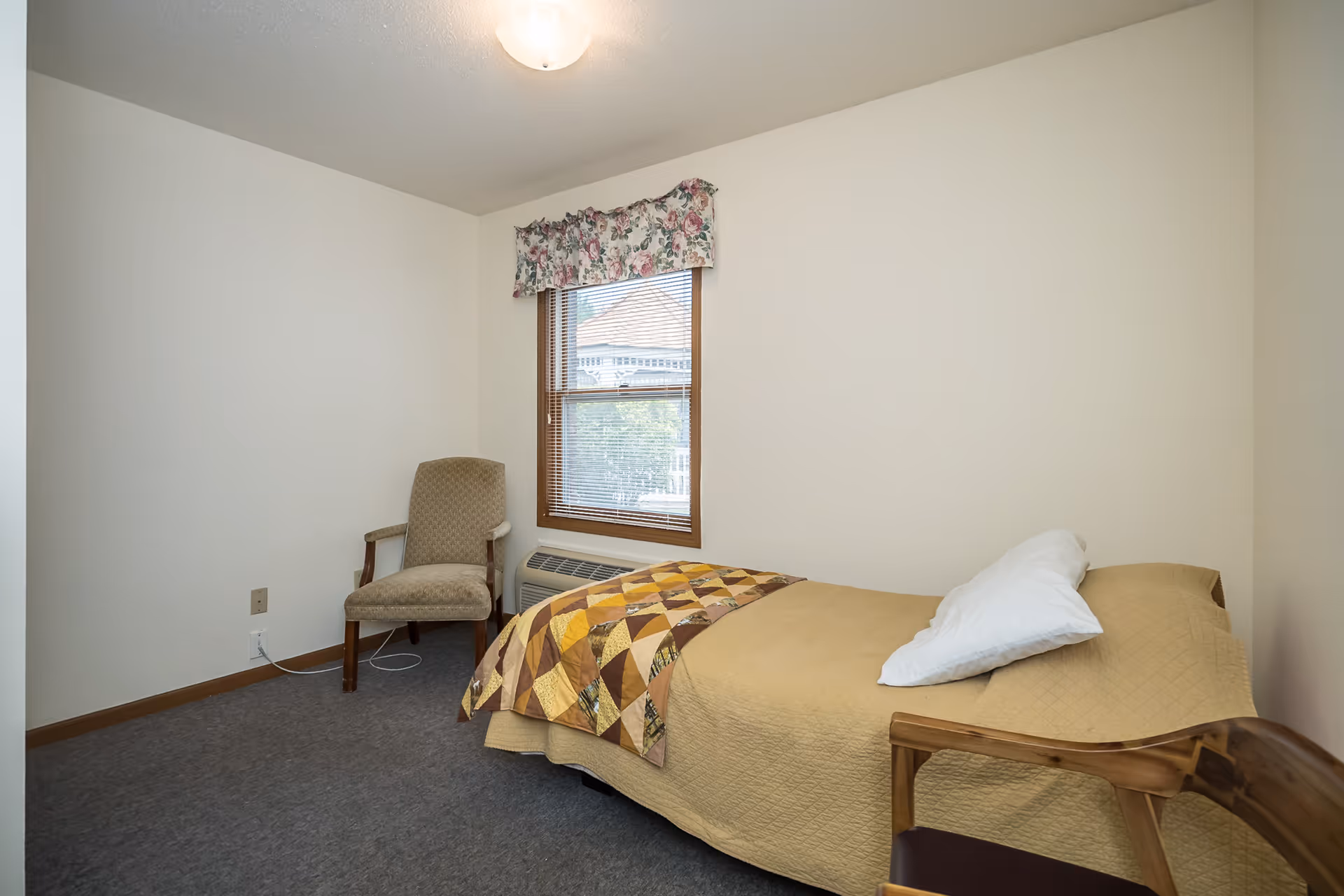 A small bedroom with a single bed covered with a beige quilt and a patchwork blanket, a white pillow, a wooden chair with a cushion, a beige upholstered armchair, a window with blinds and a floral valance, and a wall-mounted heater below the window.