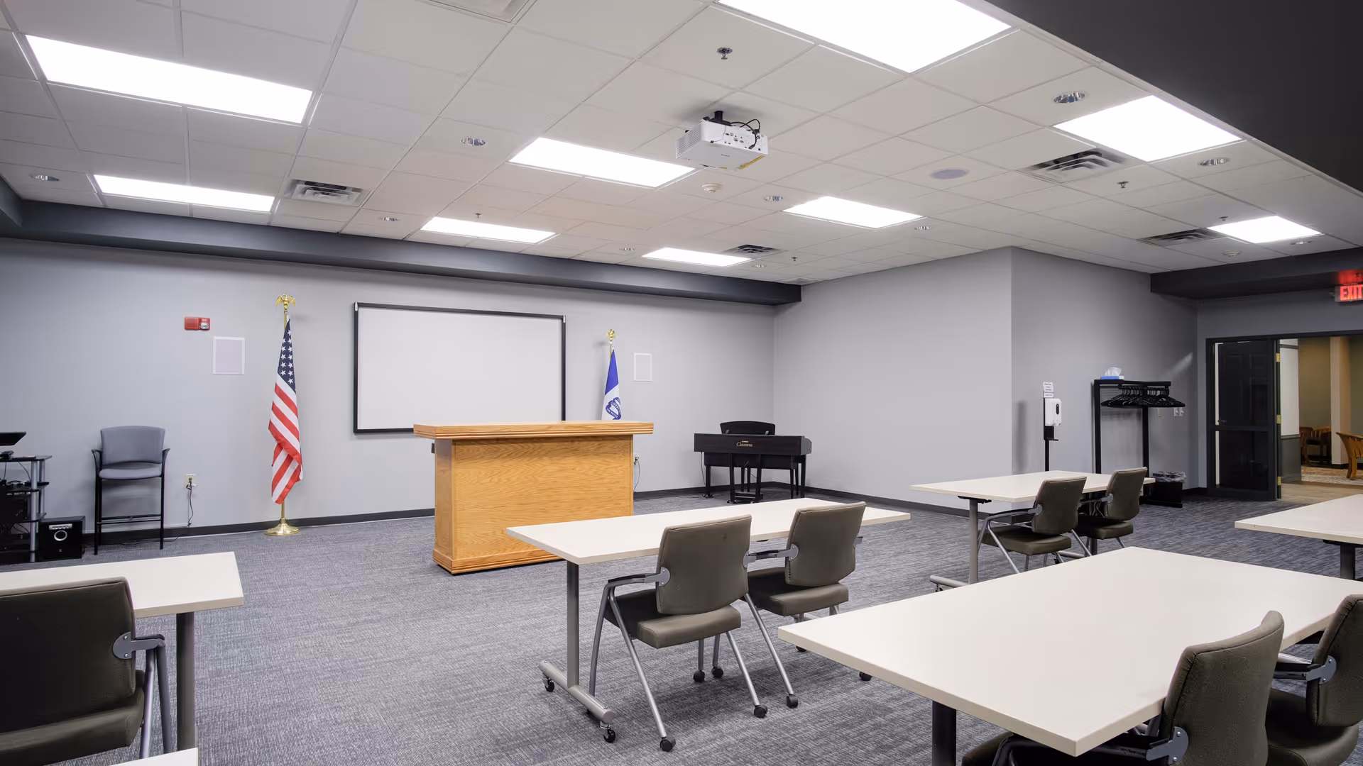 A conference or meeting room with several tables and chairs arranged facing a wooden podium and a large projection screen on the wall. The room has a gray carpet, gray walls, and ceiling lights. An American flag and another flag are positioned on either side of the screen. There is a piano in the corner and a coat rack near the entrance.