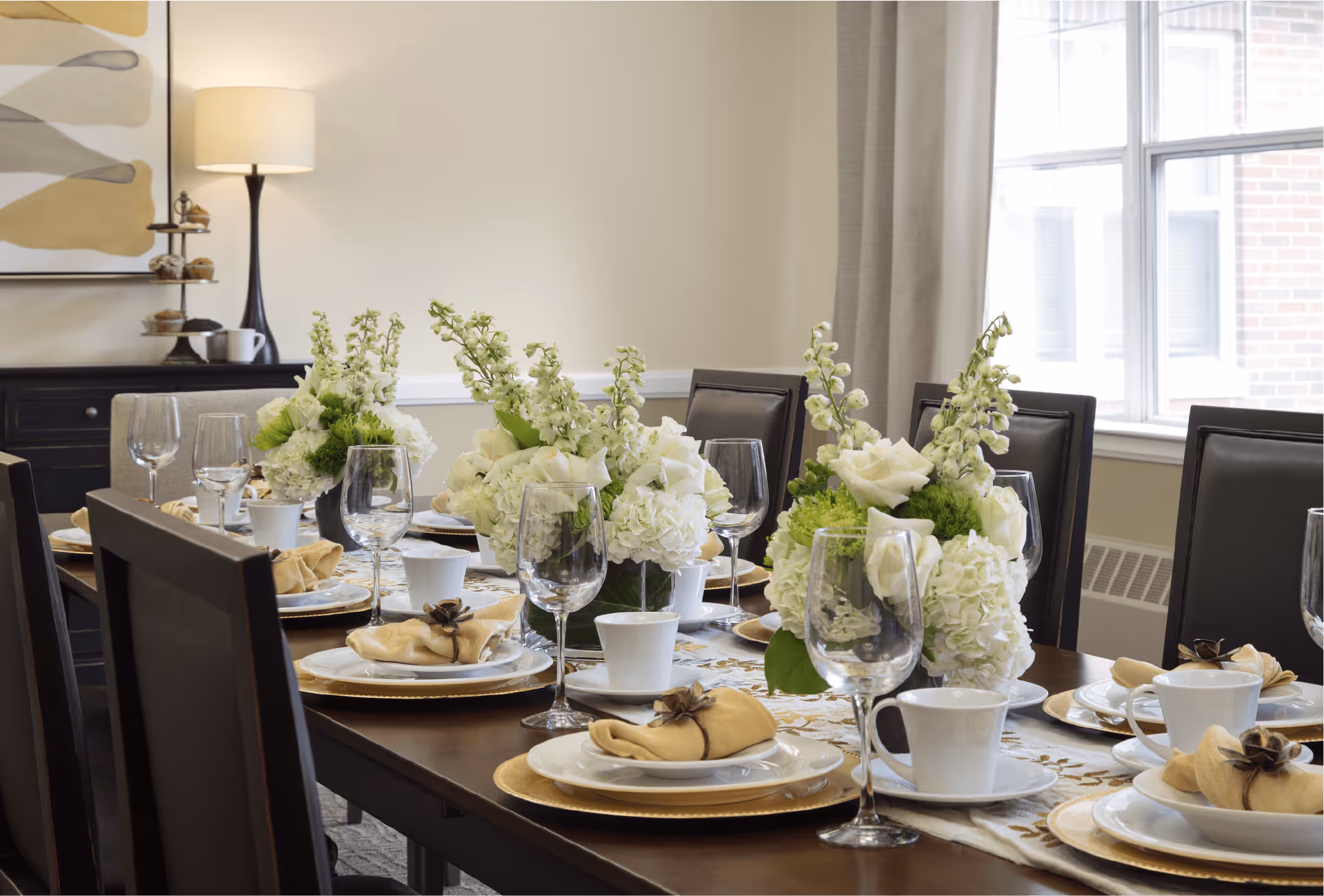 A dining table set for a meal with white plates, beige napkins, white cups, and wine glasses. The table is decorated with three floral centerpieces featuring white and green flowers. In the background, there is a sideboard with a lamp and a tiered tray with pastries, and a large window with beige curtains letting in natural light.