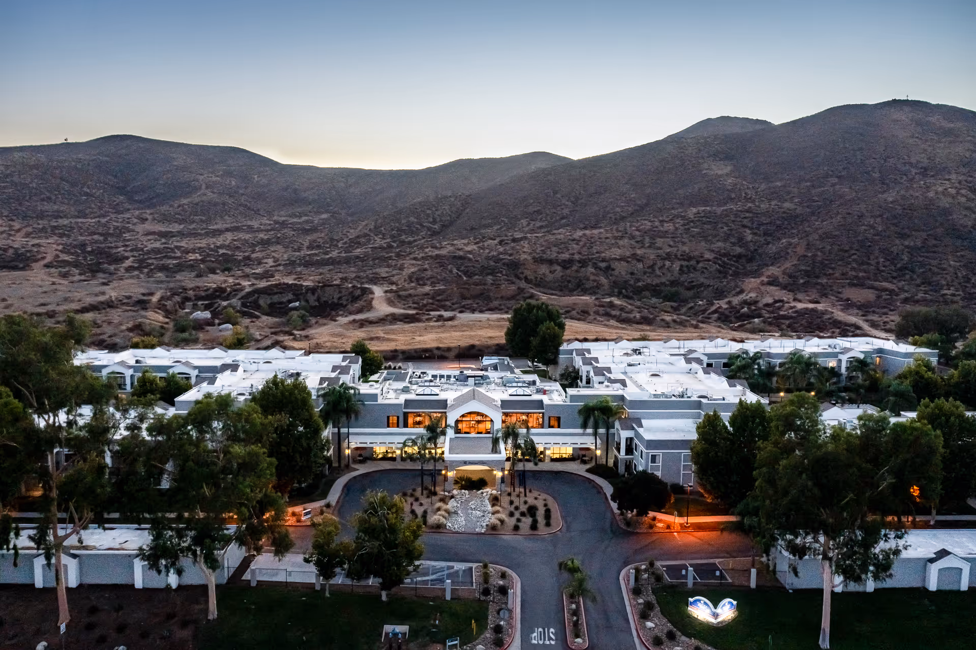 Aerial view of Menifee Senior Living facility at dusk, showing a large white building complex surrounded by trees and landscaping, with hills in the background and a circular driveway in front.