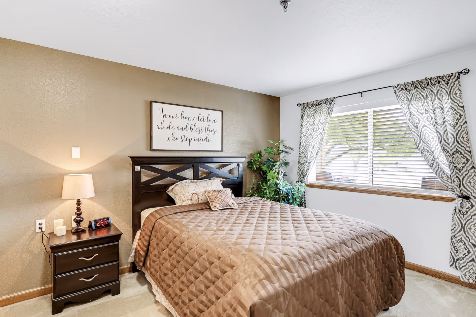 A bedroom with a neatly made bed covered with a brown quilt. There is a dark wooden headboard and a nightstand with a lamp, candles, and a digital clock showing 2:56. A framed sign above the bed reads, 'In our home let love abide and bless those who step inside.' A large window with patterned curtains lets in natural light, and a green potted plant is placed near the window.