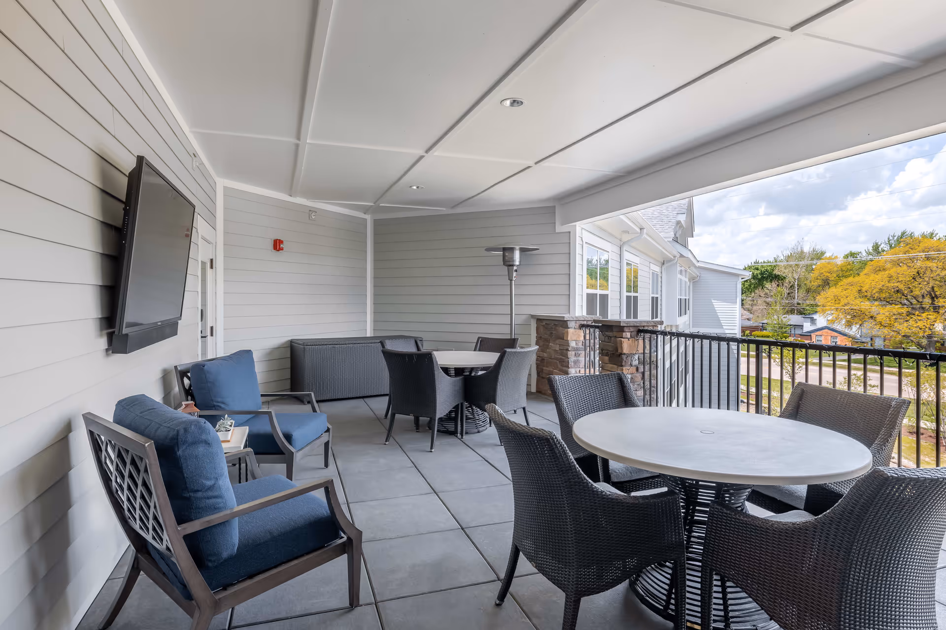 Covered outdoor patio area with gray tiled floor, gray siding walls, and a white ceiling. The space features a mounted flat-screen TV, two cushioned armchairs with blue cushions, a small side table, two round tables each surrounded by four wicker chairs, a tall outdoor heater, and a railing overlooking a street with trees and houses.