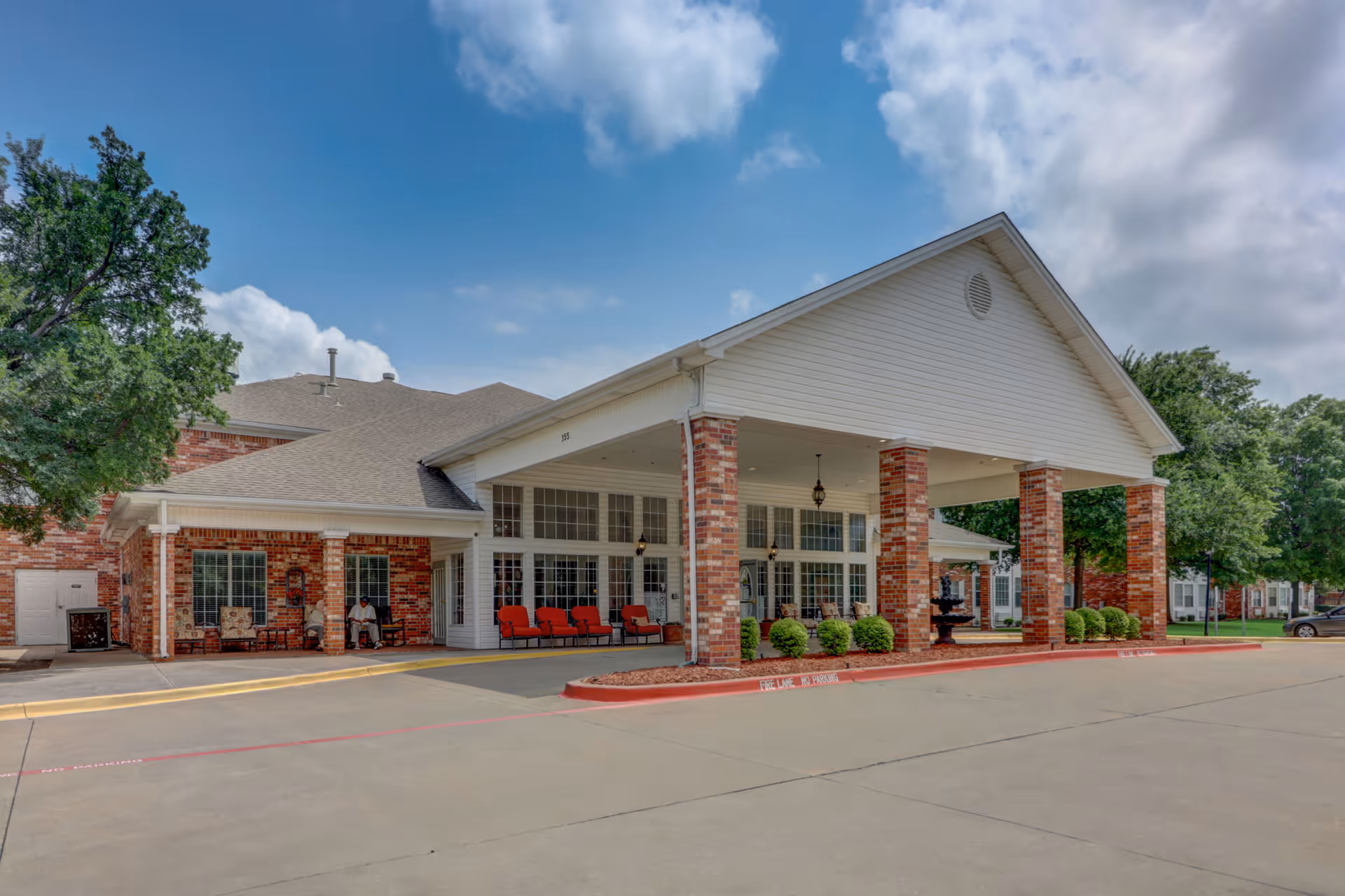 Exterior view of Morada Grand Prairie facility showing a covered entrance with brick pillars, red cushioned chairs, and a small fountain surrounded by greenery under a partly cloudy sky.