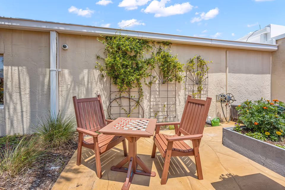 Outdoor patio area with two wooden chairs and a small table featuring a checkerboard pattern. The patio is adjacent to a beige stucco wall with climbing plants on trellises. There is a garden bed with flowers and greenery on the right side under a clear blue sky with some clouds.