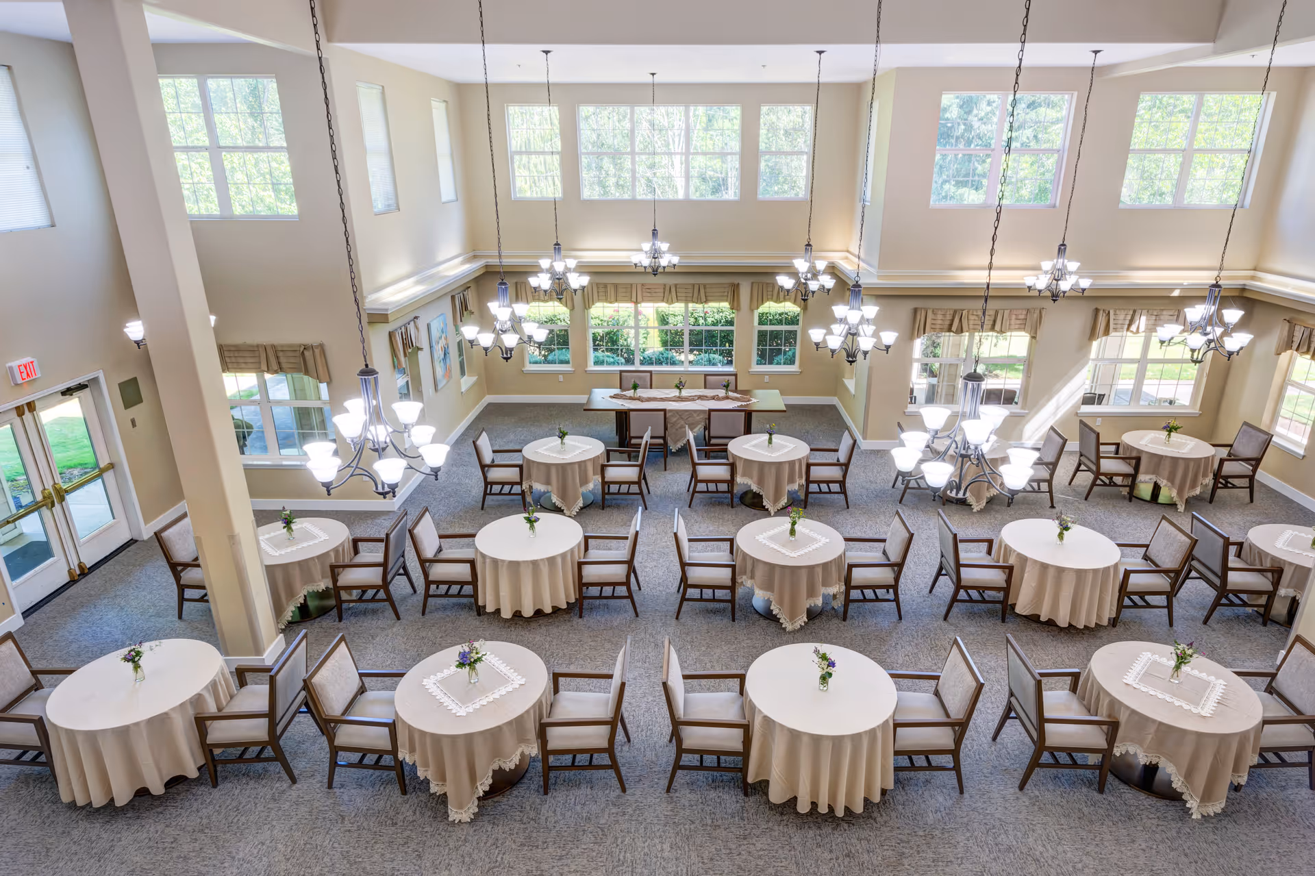 Bright and spacious dining room with multiple round tables covered with beige tablecloths, each adorned with a small flower vase. The room features large windows allowing natural light to fill the space, beige walls, and multiple elegant chandeliers hanging from the high ceiling. Chairs are neatly arranged around the tables, and there is a double door exit on the left side.
