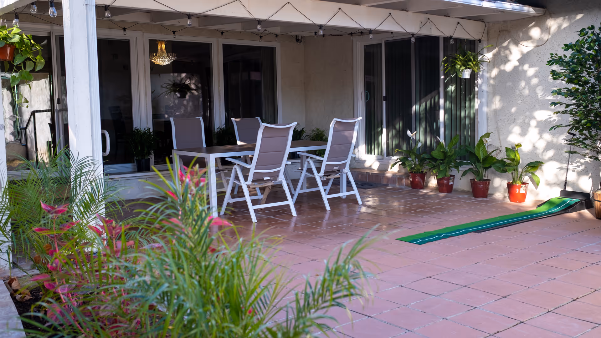 Outdoor patio area with a table and four chairs under a pergola. Several potted plants are placed along the wall and hanging from the pergola. The patio floor is tiled, and there is a green putting mat on the ground.
