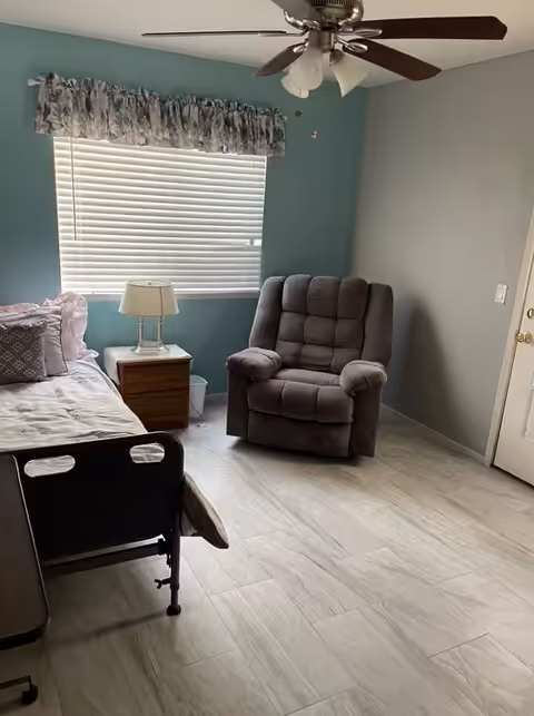 A small bedroom with a hospital-style bed on the left, a wooden nightstand with a lamp, a large window with closed blinds and a floral valance, a gray cushioned recliner chair, a ceiling fan with lights, and light-colored wood flooring.