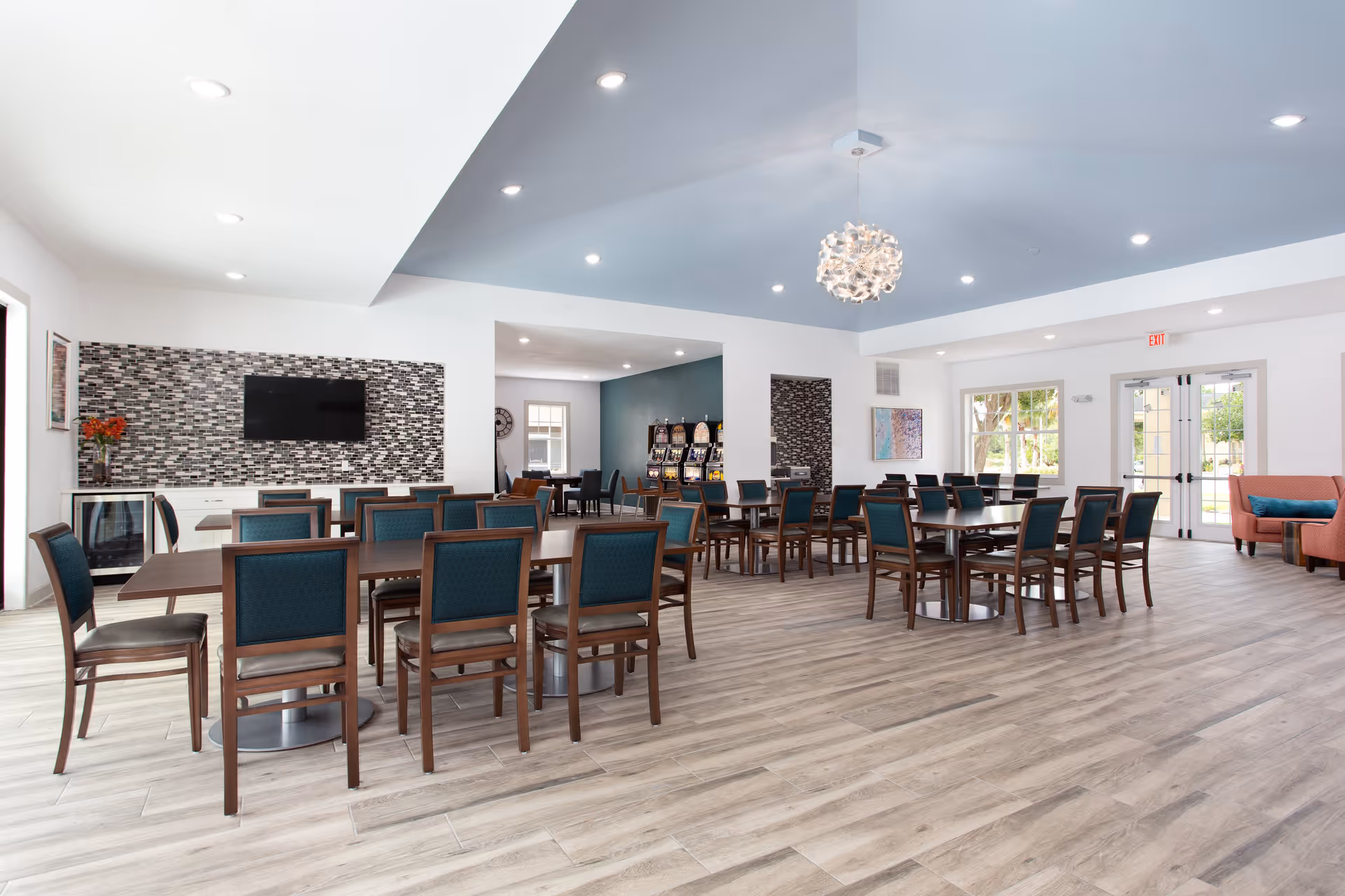 A spacious and well-lit dining room in Glenbrooke Senior Living featuring multiple wooden tables with teal cushioned chairs. The room has light wood flooring, a modern chandelier, a wall-mounted TV, and large windows and glass doors allowing natural light to enter. There is also a small seating area with a coral-colored couch and a blue pillow.