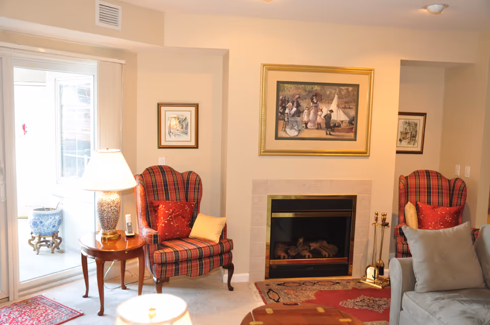 Cozy living room with two red plaid armchairs each with decorative pillows, a wooden side table with a lamp and a cordless phone, a fireplace with a tiled surround, and framed artwork above and beside the fireplace. A sliding glass door leads to an outdoor area with a blue and white planter visible.