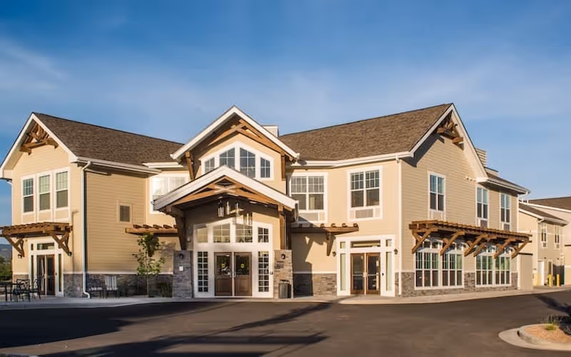 Exterior view of a two-story senior living facility building with beige siding, large windows, and wooden architectural accents under a clear blue sky.