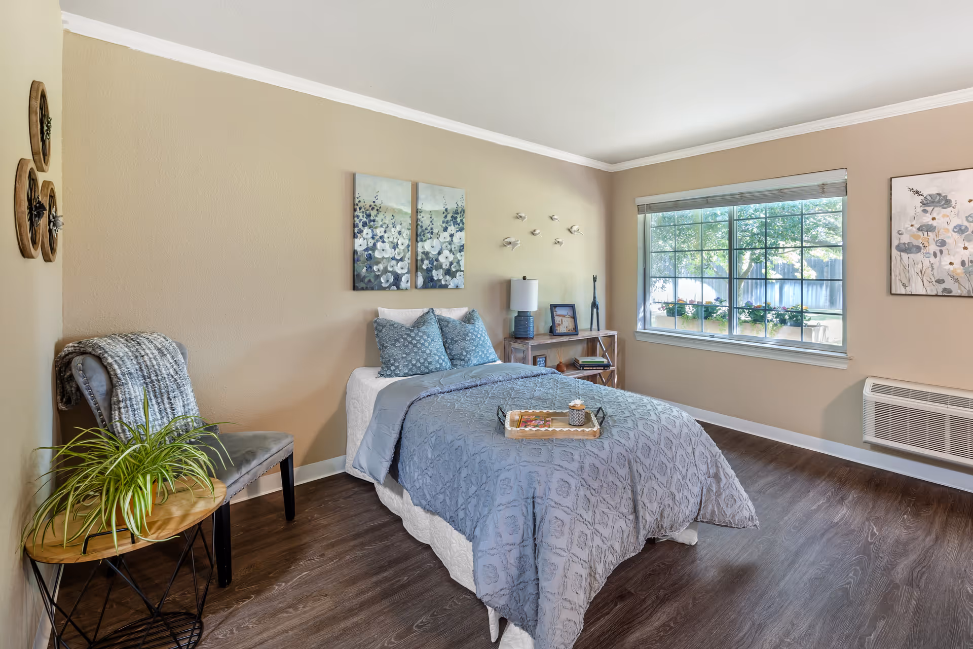 A cozy bedroom with a single bed covered in a blue quilt and matching pillows. There is a small wooden side table with a lamp, a framed picture, and decorative items. A window with blinds lets in natural light, showing greenery outside. A gray chair with a blanket draped over it and a small round table with a potted plant are also visible. The walls are beige with floral artwork and decorative wall hangings.