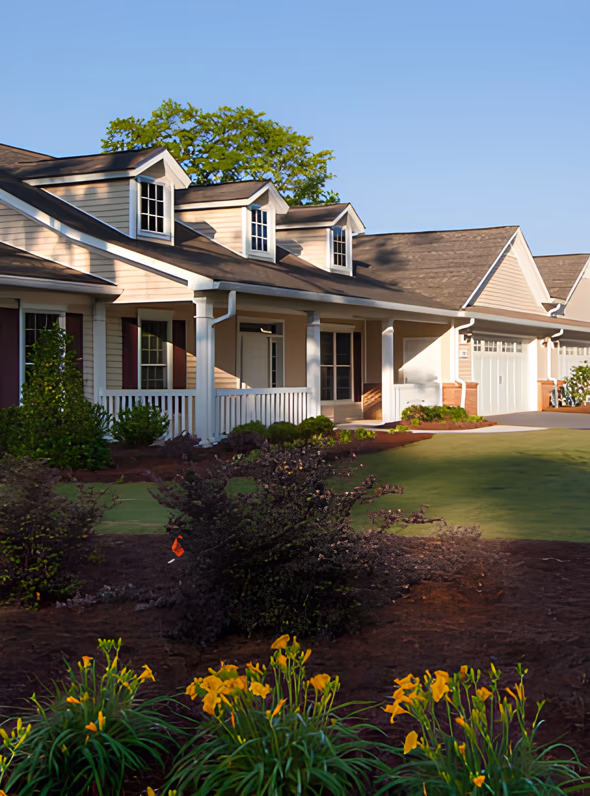 Exterior view of a residential-style building with a porch, white railings, and dormer windows on the roof. The foreground features a landscaped garden with green shrubs and yellow flowers under a clear blue sky.