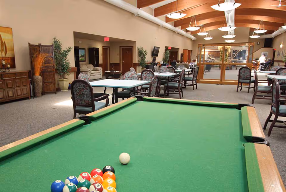 Large communal activity room with a pool table in the foreground and tables and chairs under wooden beam ceilings.
