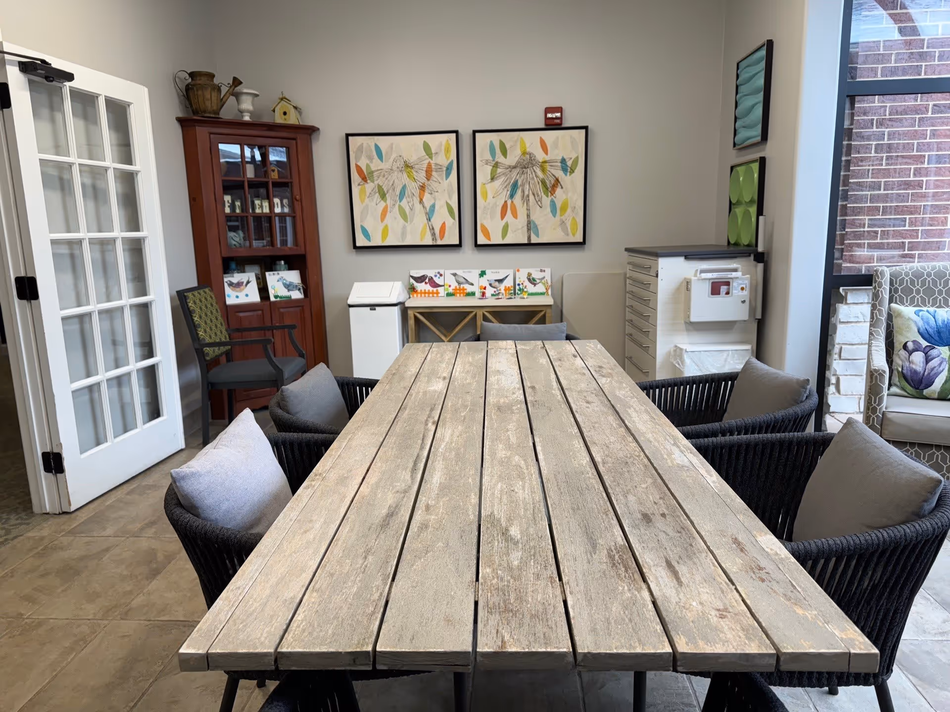 A cozy interior room with a long wooden table surrounded by six cushioned chairs. The room features a glass-paneled door on the left, a wooden cabinet with decorative items, two framed artworks of colorful leaves on the wall, and a small table with bird-themed decorations. There is also a window on the right side with a cushioned chair nearby, and a cabinet with drawers and a mounted device on the wall.