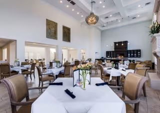 Bright, high-ceiling dining room with multiple tables set with white linens, a central chandelier, and a service counter at the back.