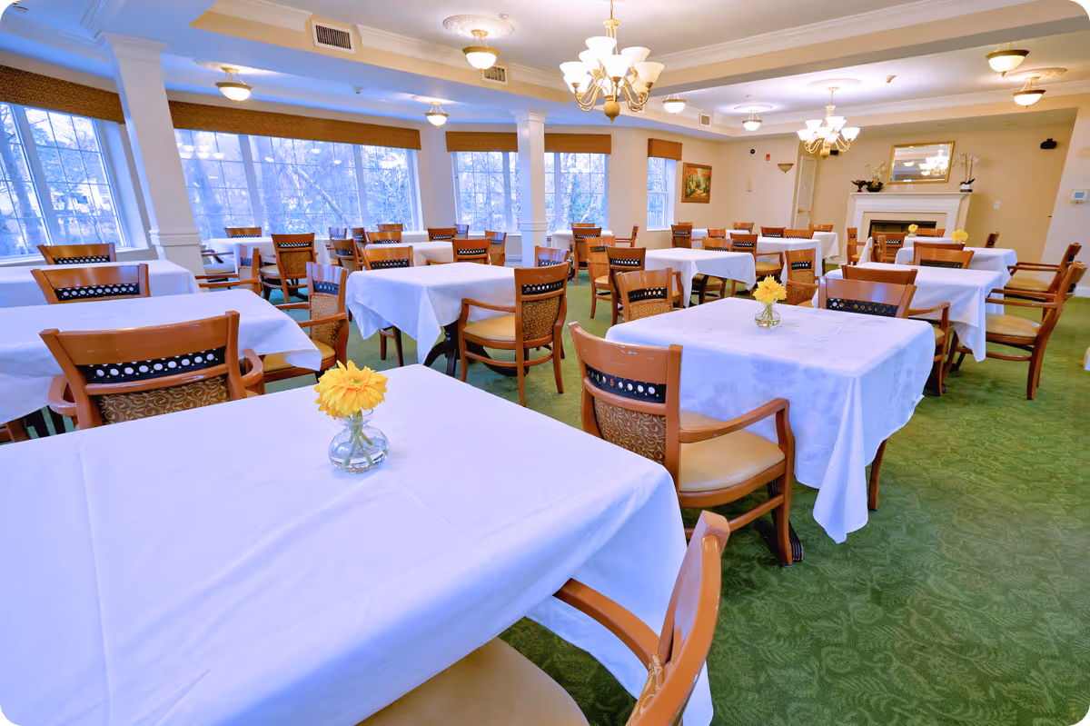 A spacious dining room with multiple tables covered in white tablecloths, each adorned with a small vase holding a yellow flower. Wooden chairs with cushioned seats surround the tables. Large windows line one side of the room, allowing natural light to fill the space. The room features green carpet, chandeliers, and a fireplace with a mirror above it.