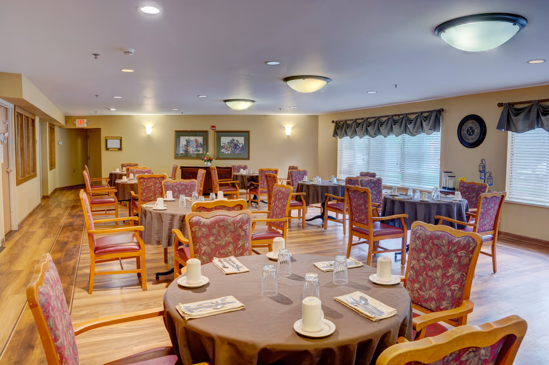 Bright dining room with round tables covered in brown tablecloths, place settings, and upholstered wooden chairs.