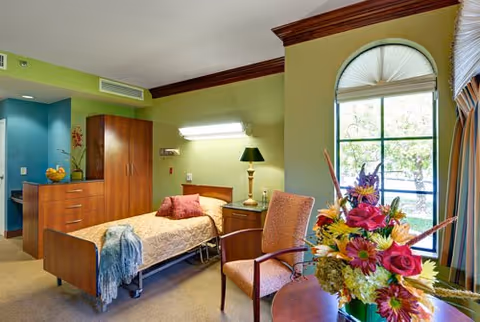 A well-lit nursing facility room with a single bed covered in a beige blanket and a blue throw. Next to the bed is a wooden nightstand with a lamp and a chair with armrests. There is a wooden cabinet and dresser against the wall, and a large window with an arched top letting in natural light. A colorful bouquet of flowers is placed on a round table near the window. The walls are painted green with dark wood trim.