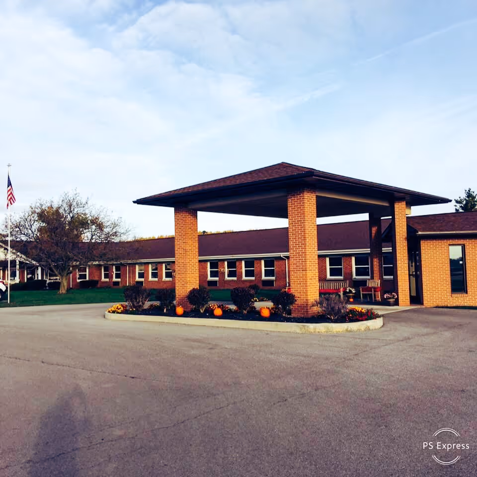 Exterior view of a single-story brick building with a covered entrance supported by four brick pillars. There are small landscaped flower beds with pumpkins and bushes around the entrance area. An American flag is visible on a flagpole to the left, and the sky is partly cloudy.