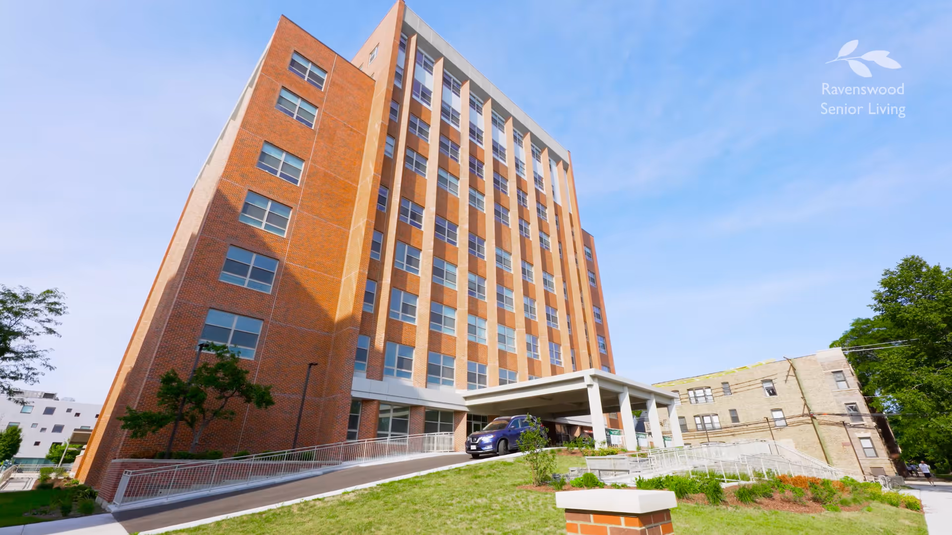 Exterior view of a multi-story brick building with many windows under a clear blue sky. There is a covered entrance with a car parked underneath and a ramp leading up to it. Green grass and some small trees surround the building. The logo and text 'Ravenswood Senior Living' are visible in the top right corner.
