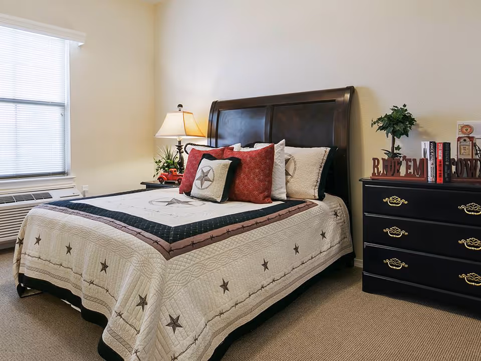 A furnished bedroom with a made bed featuring a star-patterned quilt, wooden headboard, bedside lamp and a black dresser with decorative items.