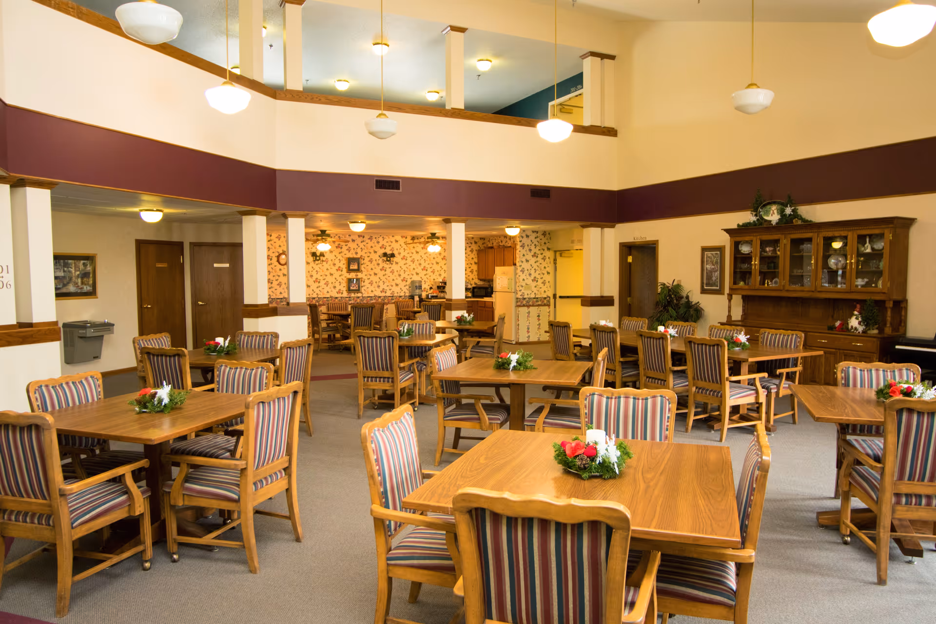 Bright communal dining room with wooden tables and striped upholstered chairs, floral centerpieces, and a high ceiling balcony.