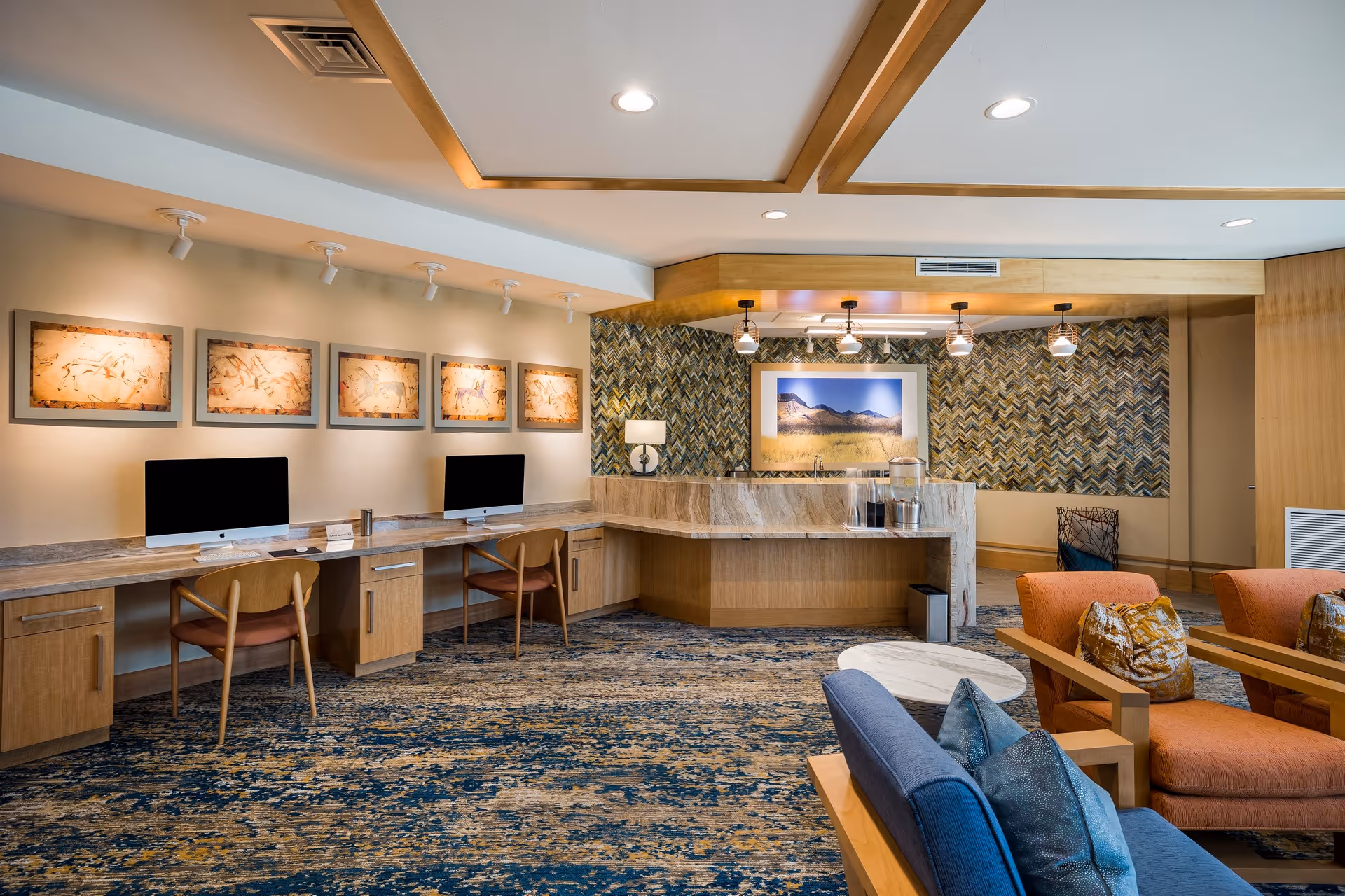 A modern common area with a long desk featuring two desktop computers and wooden chairs. The wall above the desk is decorated with five framed artworks. To the right, there is a counter with a water dispenser and a large framed photograph of a mountain landscape on a patterned wall. In the foreground, there are comfortable seating options including an orange armchair and a blue sofa with decorative pillows. The room has a patterned carpet and recessed lighting in the ceiling.