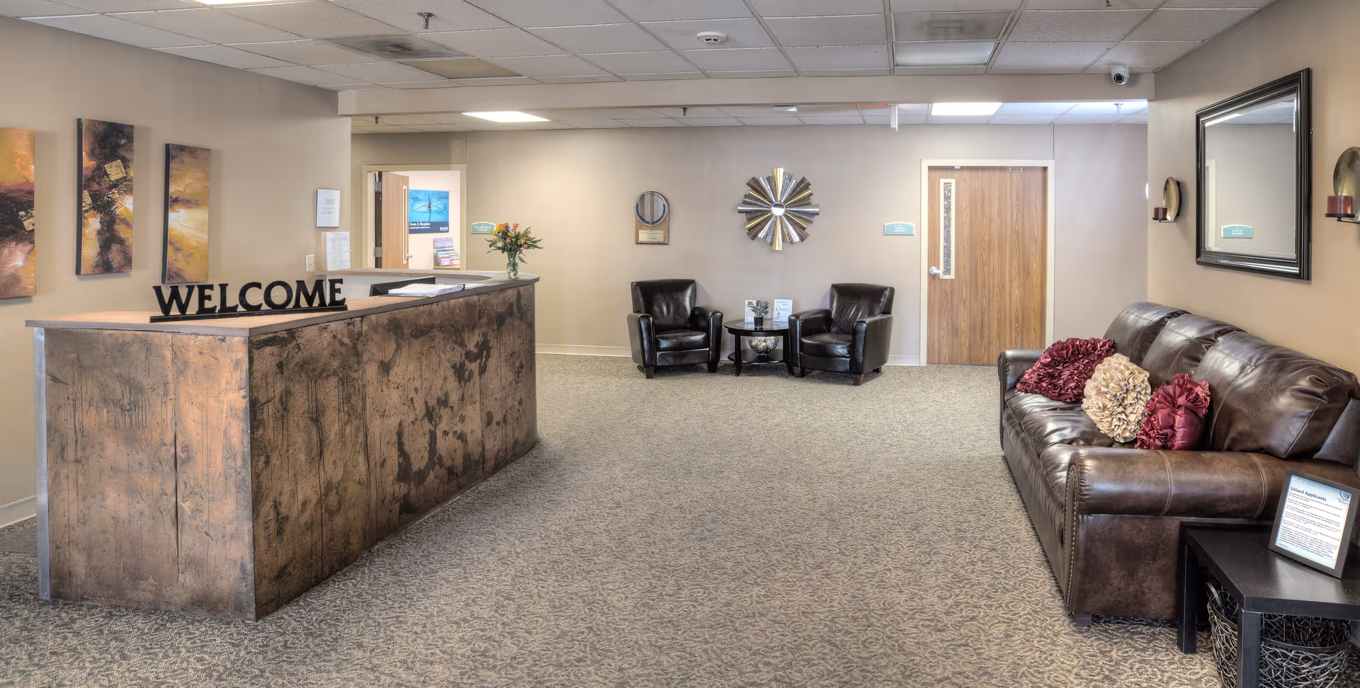 Reception area of Albuquerque Heights Healthcare and Rehabilitation Center featuring a rustic wooden welcome desk, two black leather chairs with a small table between them, a brown leather couch with decorative pillows, wall art, a large mirror, and a carpeted floor.