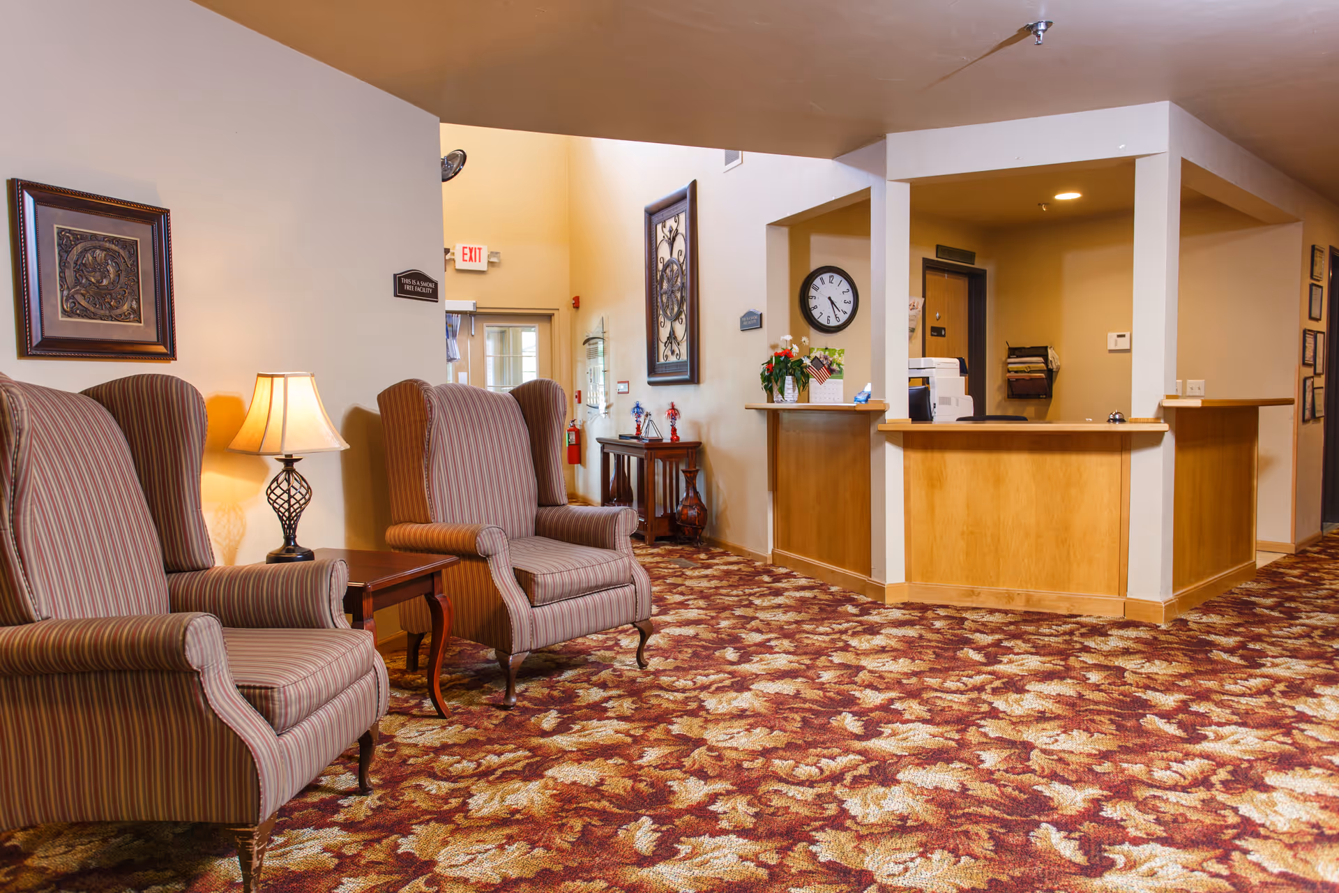 Interior view of a senior living facility reception area with a wooden front desk, two striped upholstered armchairs, a side table with a lamp, framed artwork on the walls, a clock, and a patterned carpet with autumn leaf designs.