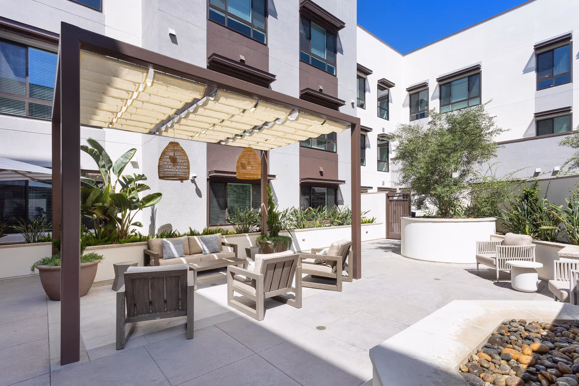 Outdoor courtyard area at a senior living facility with a pergola providing shade over a seating area consisting of a sofa and several armchairs. There are potted plants, greenery along the walls, and a fire pit with stones in the foreground. The building surrounding the courtyard has multiple windows.