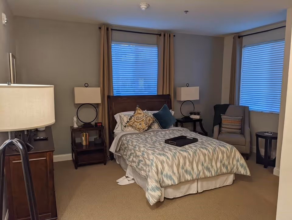 A cozy bedroom with a double bed featuring patterned bedding and decorative pillows. The room has two windows with closed blinds and beige curtains. There are two bedside tables with matching lamps, a cushioned armchair with a throw blanket and pillow, a small round side table, and a wooden dresser with a lamp on it. The walls are painted light gray and the floor is carpeted.